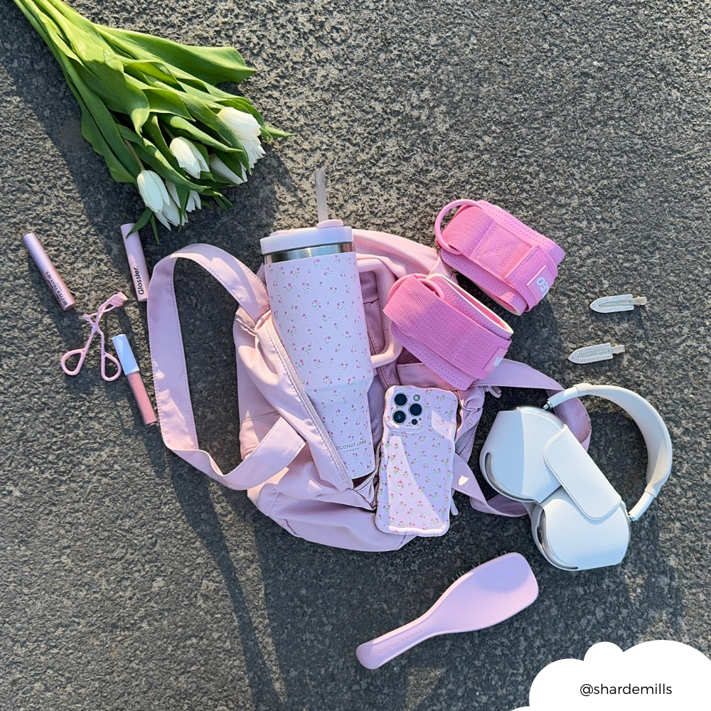Pink gym bag, pink accessories, white headphones, tulips, and pink ditsy floral wavy phone case on pavement.