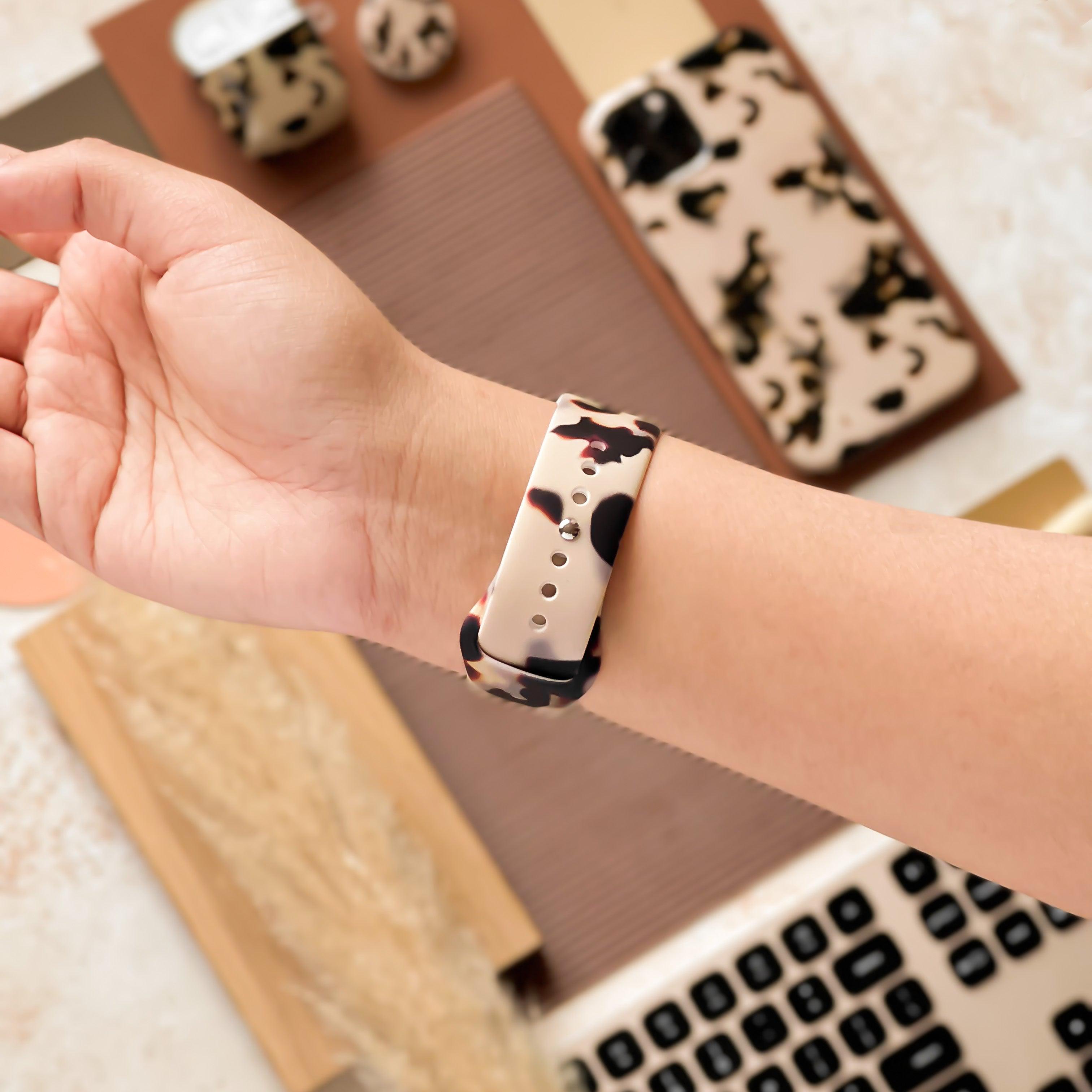 A hand wearing a smartwatch with a Milky Ivory Tort Apple Watch Strap by Coconut Lane. Surrounding the wrist are various tech accessories, including a camo phone case, wireless earbuds, and a laptop keyboard, all placed on a beige and brown textured surface.