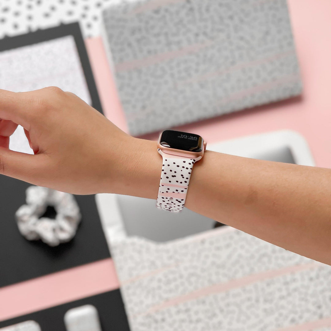 Close-up of a person's wrist wearing a smartwatch with a white band featuring black speckles. In the background are various coordinating items, including a scrunchie, notebooks, and electronic devices, in black, white, and pink tones.