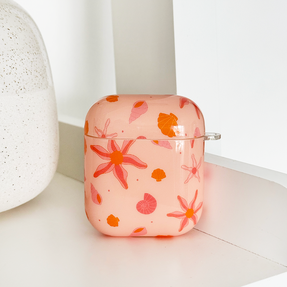 Pink floral-patterned wireless earbuds case with earbuds inside, resting against tan pampas grass on a beige background.