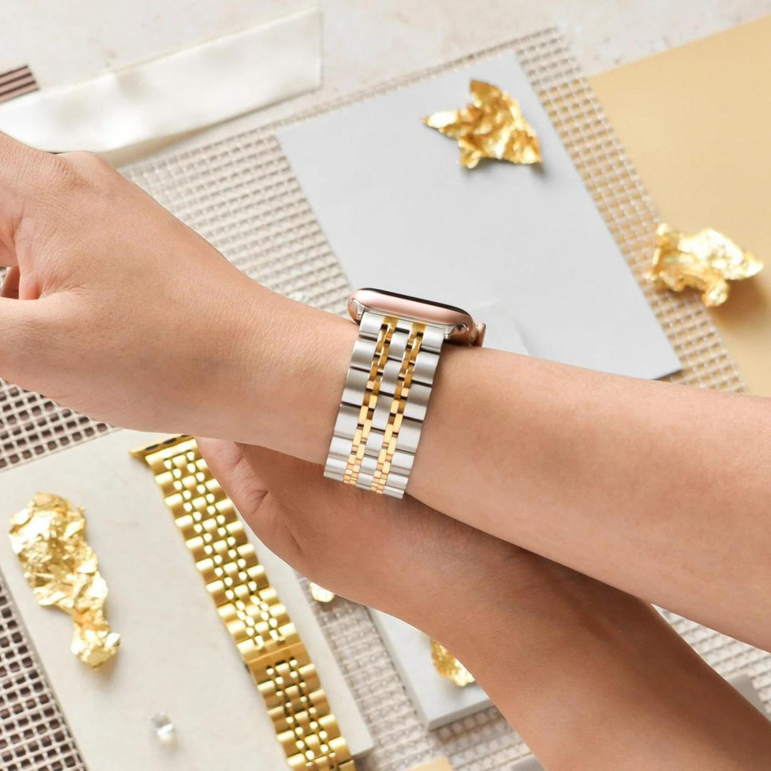 A person wearing a smartwatch with a silver and gold metal wristband. The background features various textured surfaces and pieces of gold foil.