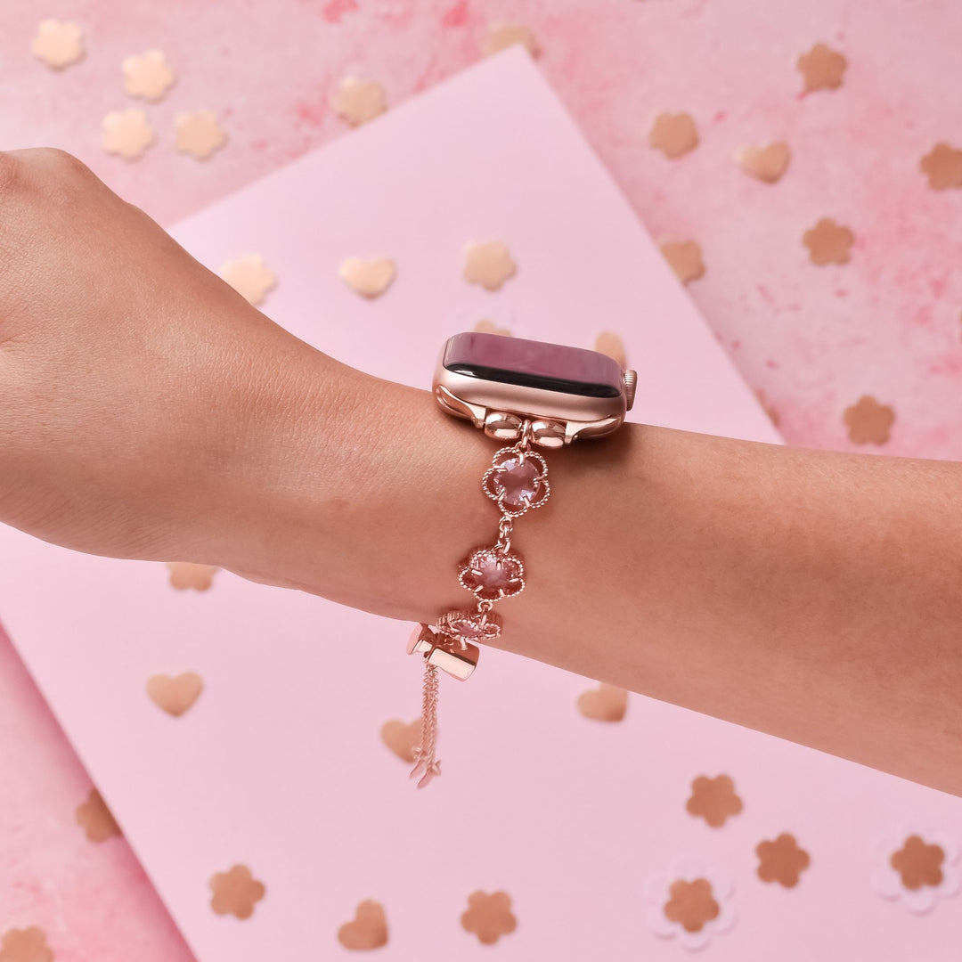 A wrist adorned with a rose gold smartwatch featuring a pink screen, and a delicate bracelet with pink flower accents. The background is pink with tiny heart and flower-shaped confetti scattered around.