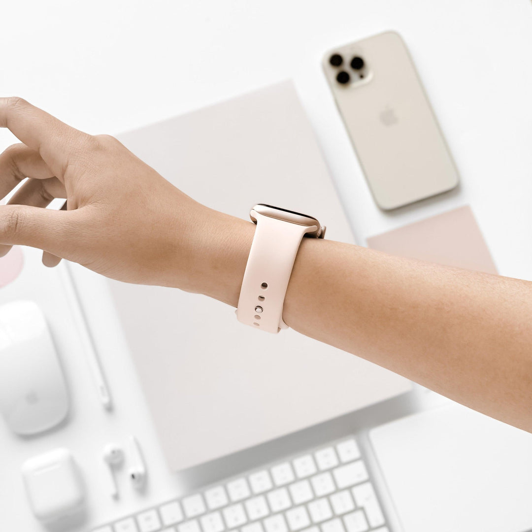 A person wearing a smartwatch with a white band on their left wrist, reaching over a white workspace with a smartphone, laptop, mouse, and earphones neatly arranged on a white surface.