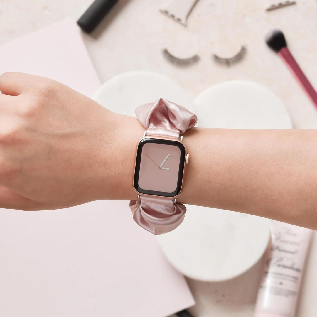 A person’s wrist wearing a smartwatch with a pink fabric band. The watch face displays a simple clock with two hands. Nearby are makeup items, including a brush, false eyelashes, and a cylindrical tube on a pale surface.