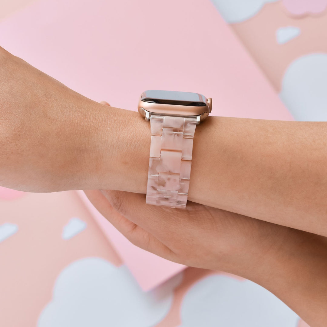 A person wearing a smartwatch with a pale pink, marble-patterned band. The background consists of soft pink and white elements, some resembling paper clouds. The person is viewed from mid-forearm to hand.