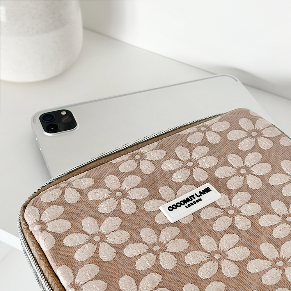 A tablet partially covered by a floral-patterned case lies on a white surface. The case features white flowers on a light brown background and a label reading "COCONUT LANE London." A ceramic pot is in the background.