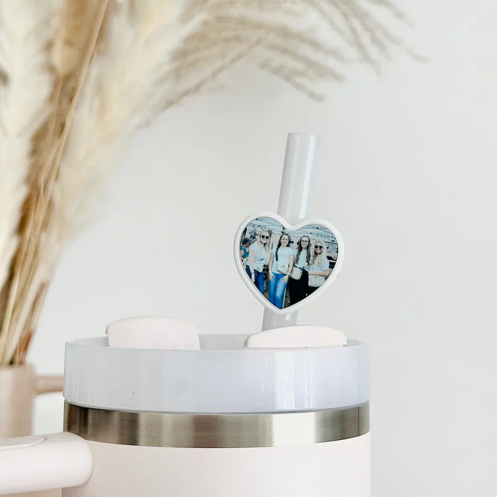 A heart-shaped photo charm of five people decorates a straw in a white tumbler, with pampas grass in the blurred background.