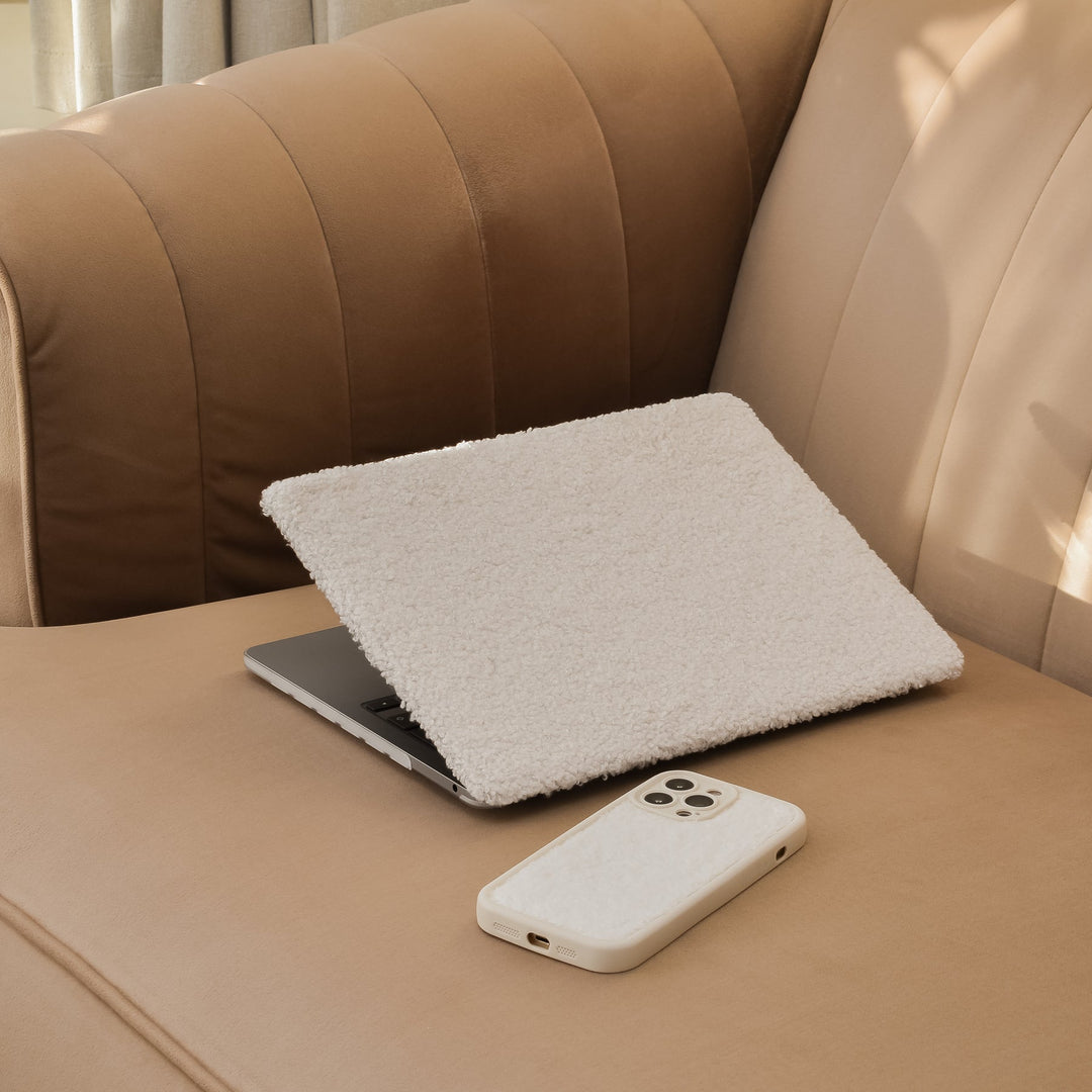A laptop with a fluffy white cover sits open on a beige couch. A smartphone with a matching fluffy white case lies next to it on the couch seat. Sunlight casts soft shadows on the couch.