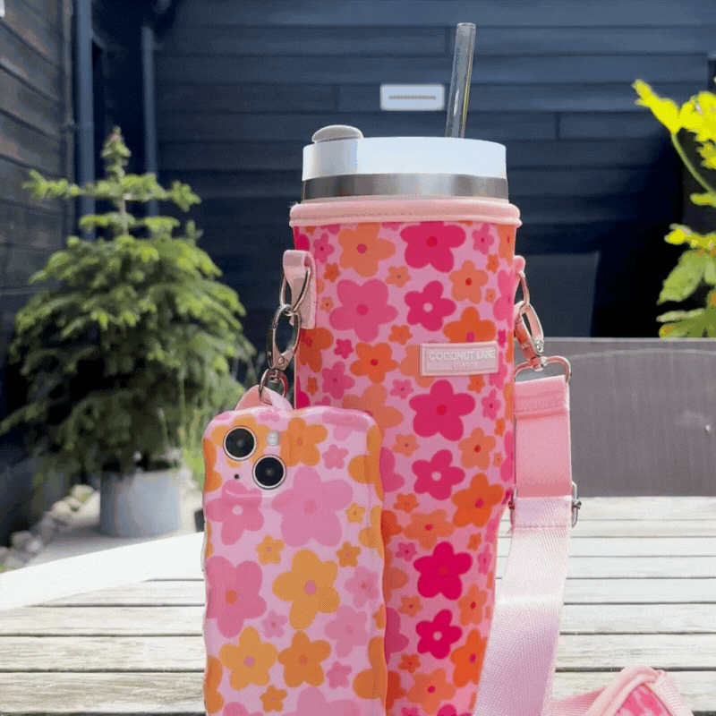 A smartphone and a water bottle, both in pink floral cases, are placed on a table outdoors. The smartphone case has a pale pink background with red and pink flowers, while the water bottle is encased in the Coconut Lane Tumbler Carry Case - Pink & Orange Bloom, featuring a similar floral pattern and shoulder strap. Behind them, greenery and a dark fence are visible.