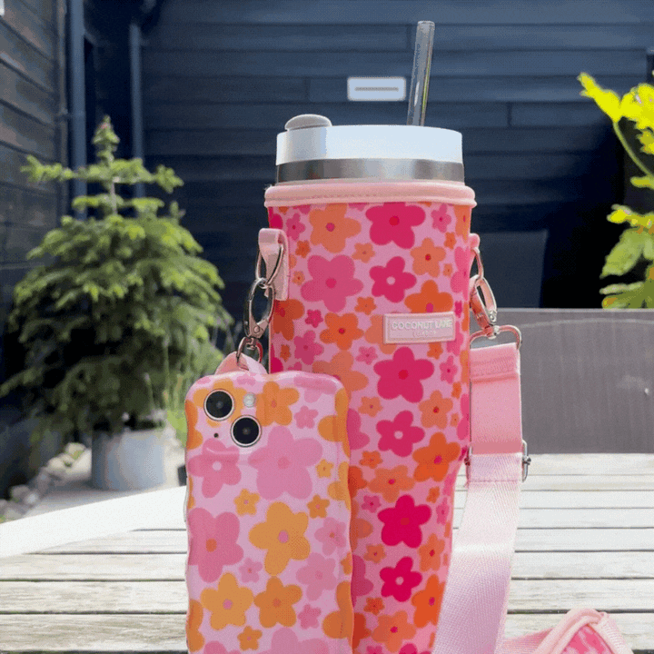 A smartphone and a water bottle, both in pink floral cases, are placed on a table outdoors. The smartphone case has a pale pink background with red and pink flowers, while the water bottle is encased in the Coconut Lane Tumbler Carry Case - Pink & Orange Bloom, featuring a similar floral pattern and shoulder strap. Behind them, greenery and a dark fence are visible.