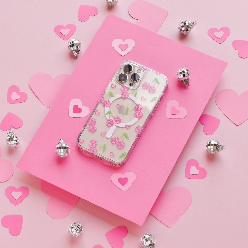 A Coconut Lane Clear MagSafe® Phone Case - Disco Cherries is placed on a hot pink rectangle surrounded by pink and light pink paper hearts. Small silver spherical decorations are scattered around, enhancing the romantic, festive vibe on a light pink background.