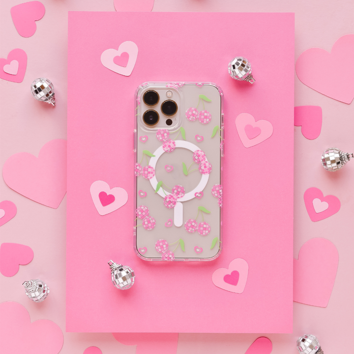 A smartphone with a Coconut Lane Clear MagSafe® Phone Case in Disco Cherries design is placed on pink paper with heart cutouts. Silver disco ball ornaments are scattered around, enhancing the playful and romantic theme.