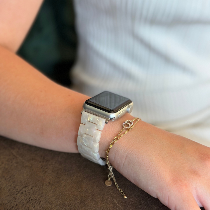 A close-up of an arm resting on a tabletop, adorned with the Luxe Iridescent Apple Watch Strap by Coconut Lane. Next to the watch is a delicate gold chain bracelet featuring a small charm. The person is wearing a white, sleeveless top.