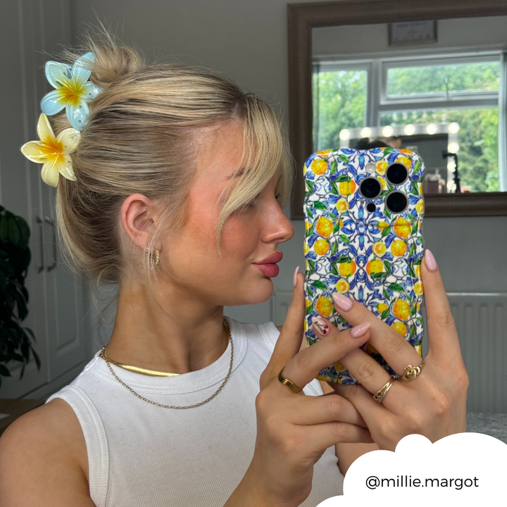 A blonde woman takes a mirror selfie with her phone adorned in the Wavy Phone Case - Amalfi Lemon by Coconut Lane. She wears a white top, gold hoop earrings, and necklaces. Her hair is tied up with large floral hair clips. The background includes a window and home interior. Instagram handle @millie.margot.