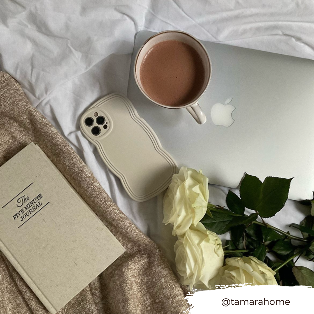 A cozy scene featuring an open MacBook with a cup of coffee on top, a beige iPhone with a highly protective NAKD Curvy Phone Case - Vanilla by Coconut Lane, a book titled "The Five-Minute Journal," and six white roses. In the corner, a handle with the username @tamarahome is displayed on a brushstroke-style background.