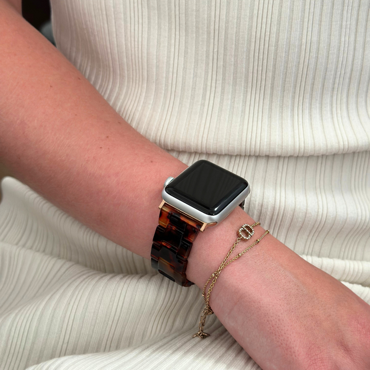 A person wearing the Luxe Tortoiseshell Apple Watch Strap by Coconut Lane on their wrist, paired with a delicate gold bracelet featuring a small charm. The person is dressed in a white, ribbed sleeveless top.