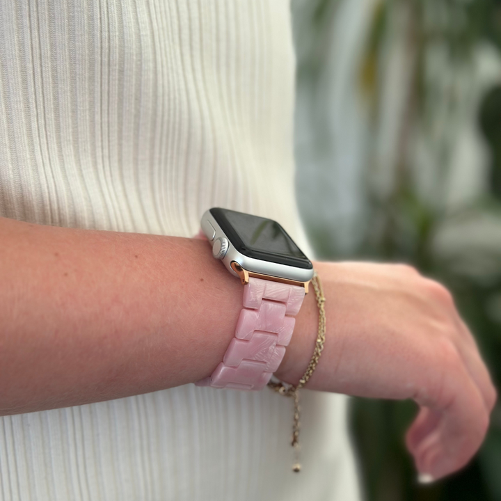 A person wearing a white ribbed top is displaying their wrist, adorned with a Luxe Pink Apple Watch Strap from Coconut Lane. Their wrist also features a delicate chain bracelet with a small charm. The background is softly blurred, revealing hints of green foliage.