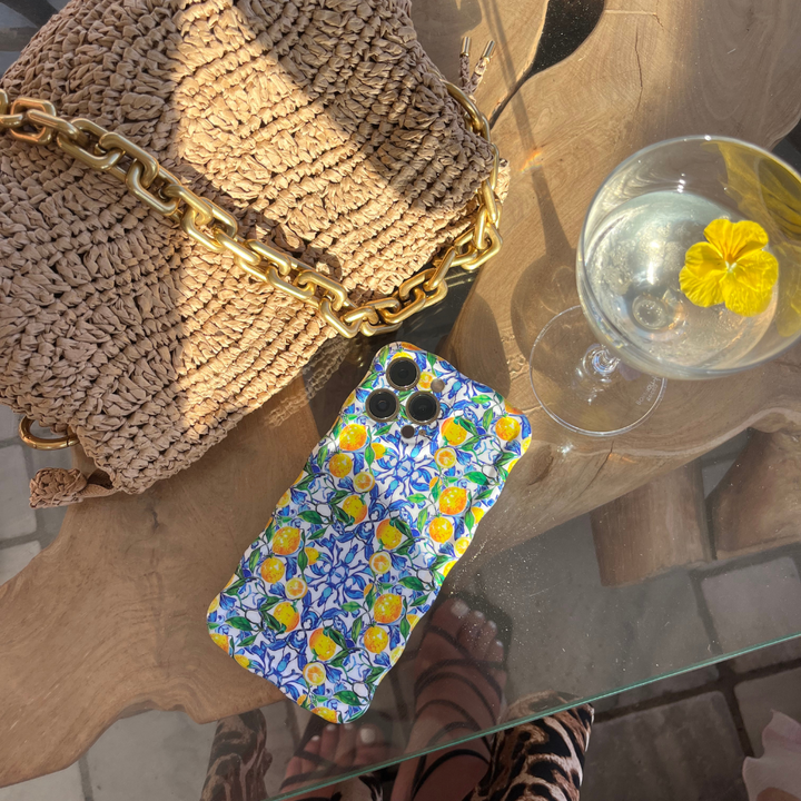 A woven straw handbag with a chunky gold chain strap is placed on a wooden table. Next to it is a phone with Coconut Lane's Wavy Phone Case in Amalfi Lemon. A glass with a clear beverage and a yellow flower floating on top is nearby. The table rests on a glass surface.