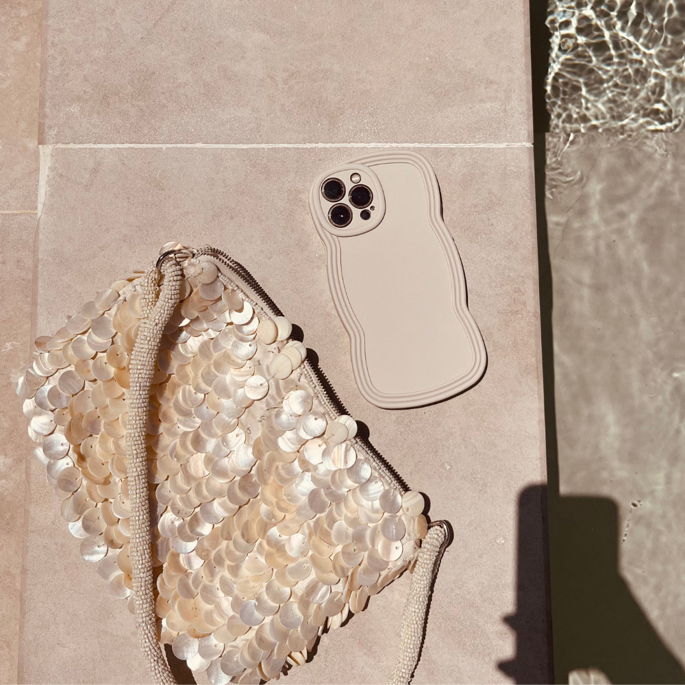 A beige smartphone with a wavy-edged NAKD Curvy Phone Case - Vanilla by Coconut Lane lies next to a shimmery bag adorned with large, circular sequins. Both lightweight items are on a light-colored surface near a pool, with ripples in the water reflecting sunlight. A shadow is partially visible in the bottom right corner.