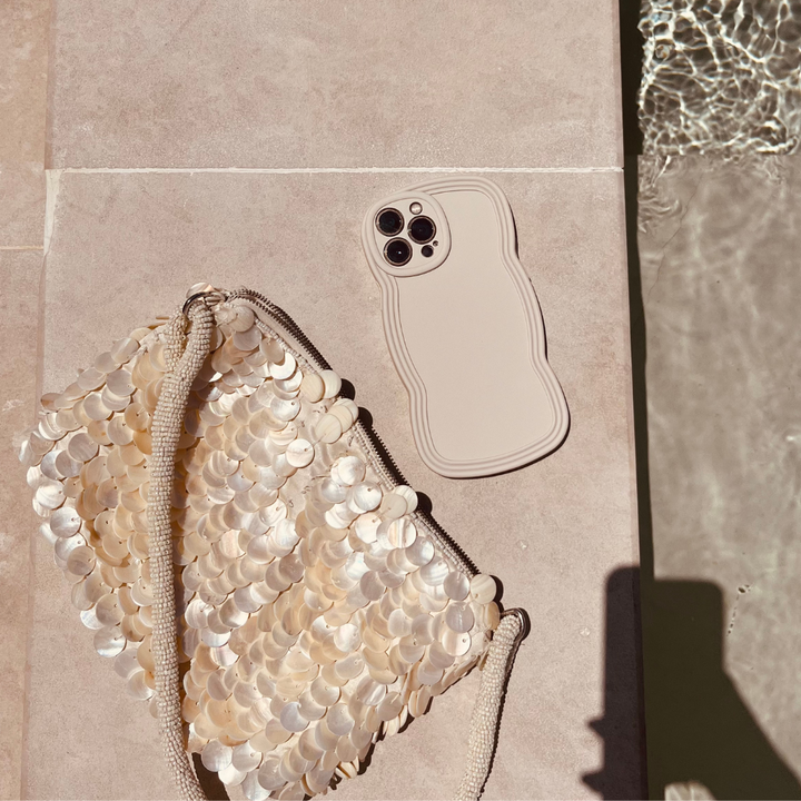 A beige smartphone with a wavy-edged NAKD Curvy Phone Case - Vanilla by Coconut Lane lies next to a shimmery bag adorned with large, circular sequins. Both lightweight items are on a light-colored surface near a pool, with ripples in the water reflecting sunlight. A shadow is partially visible in the bottom right corner.