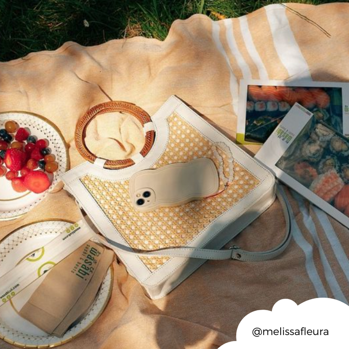 A picturesque picnic setup featuring a woven tote bag, a smartphone encased in the highly protective NAKD Curvy Phone Case - Vanilla by Coconut Lane, a plate of fruit, a small round loaf of bread, and magazines laid out on a blanket in the grass. The image is warm and sunny, depicting a relaxing outdoor scene. (@melissafleura).