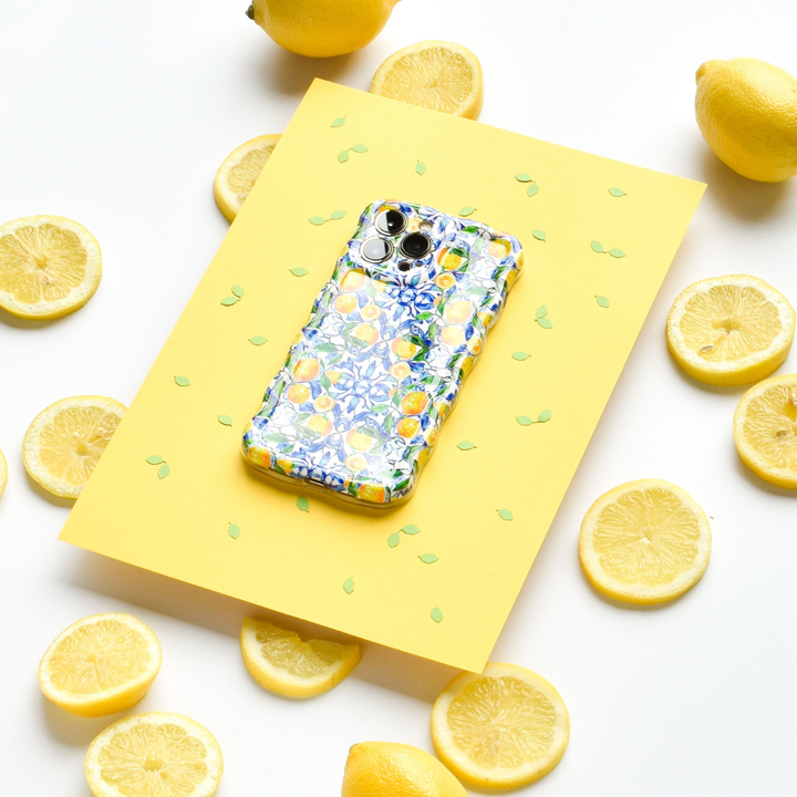 A Coconut Lane Wavy Phone Case - Amalfi Lemon sits on a yellow sheet of paper decorated with green leaf patterns. Surrounding the paper are whole and sliced lemons, creating a bright and cheerful scene.