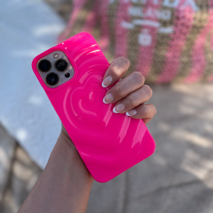 A hand with manicured nails holds the Coconut Lane Melting Heart Phone Case in bright pink, showcasing its extra protective design and 3D melting heart effect on the back. The background is slightly blurred, revealing a patterned bag and some outdoor furniture.