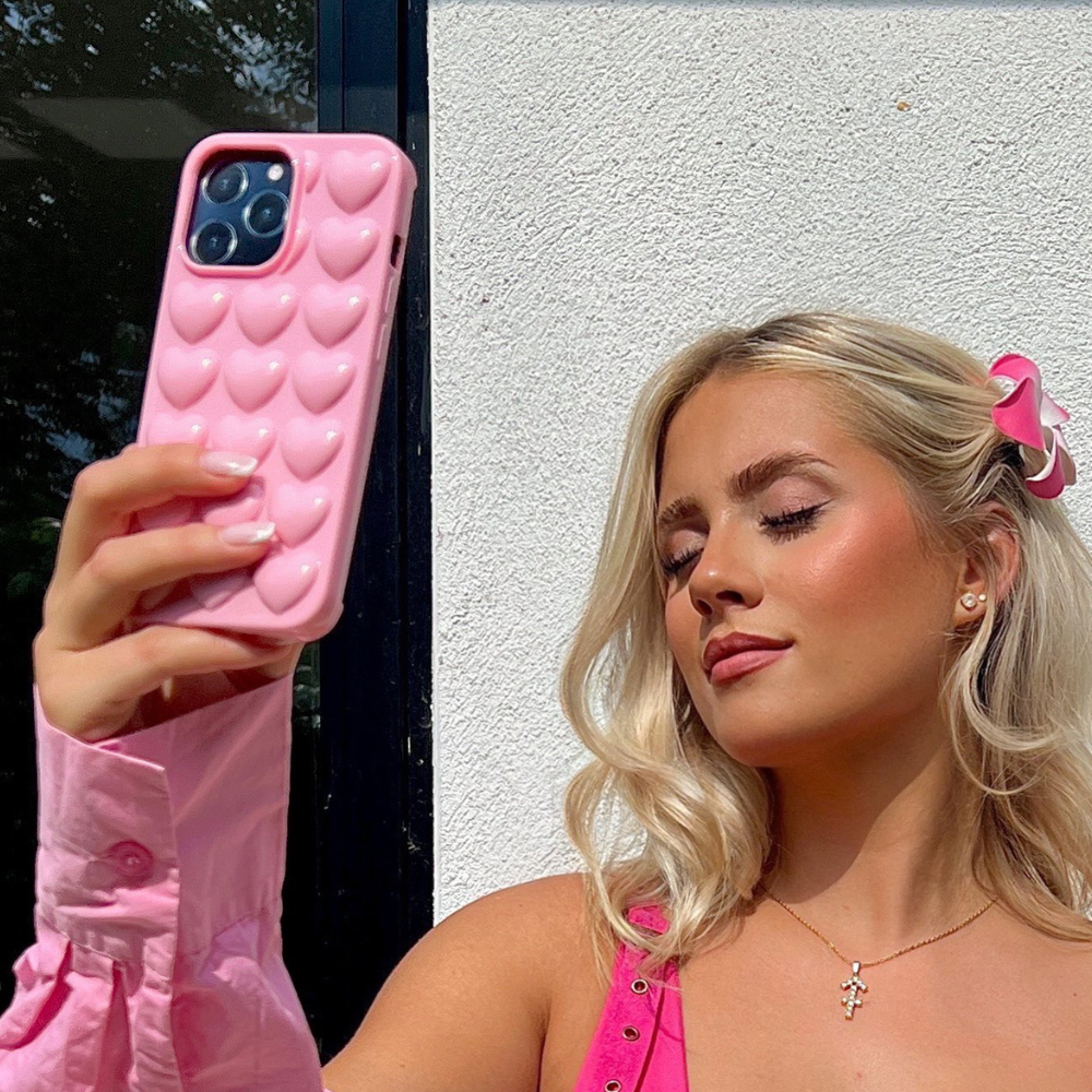A woman with blonde hair holds up her Coconut Lane 3D Hearts Phone Case in Candyfloss Pink as she takes a selfie. She is wearing a pink top, a gold necklace with a cross pendant, and a matching pink hair clip. The background displays a white wall and the edge of a window.