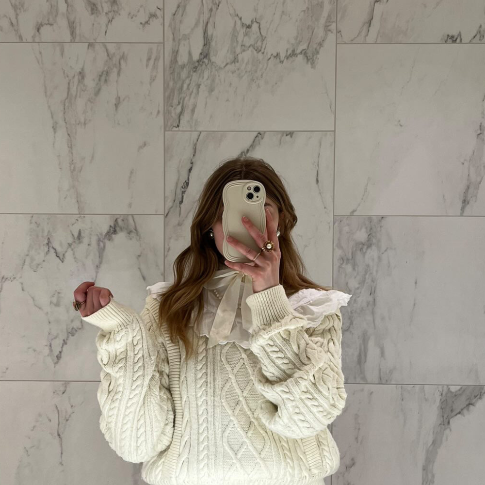 A person stands in front of a marble wall, taking a mirror selfie with a phone adorned in the highly protective NAKD Curvy Phone Case - Vanilla by Coconut Lane. They are wearing a cream-colored cable knit sweater with a white ruffled collar and lace details. Only their torso and part of their head are visible, with the phone covering their face.