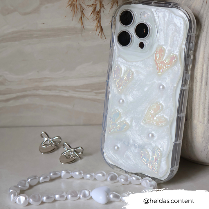 A smartphone with a pearlescent case featuring heart designs and small pearls is propped up, adorned with Coconut Lane's Beaded Phone Strap - Chunky Pearls. Silver butterfly earrings and a bracelet with white heart-shaped and spherical beads are placed beside it. The Instagram handle @heldas.content is visible in the bottom-right corner.