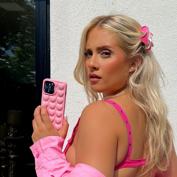 A blonde woman wearing a candyfloss pink outfit and a hair clip is holding Coconut Lane’s 3D Hearts Phone Case in Candyfloss Pink. She poses outdoors in front of a white wall with a window beside her, looking towards the camera with a serious expression.