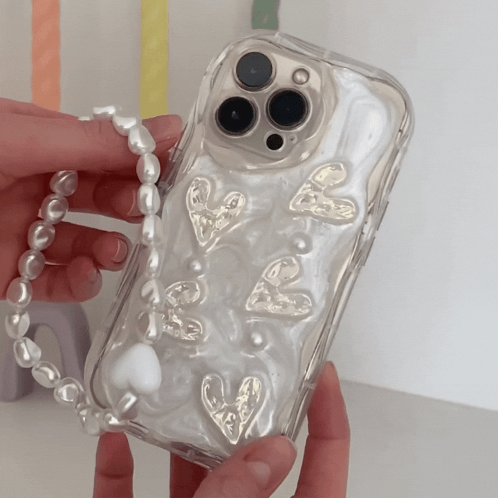 A person is holding a smartphone with a pearly white case adorned with shiny golden heart designs. Attached to the case is Coconut Lane's Beaded Phone Strap - Chunky Pearls, adding an elegant touch. The background is out of focus, showcasing some pastel-colored objects.