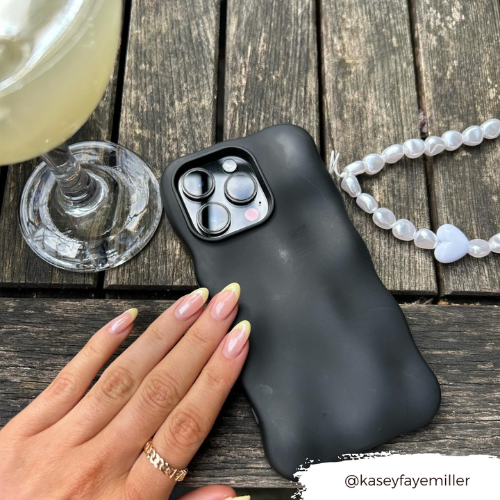 A hand with a French manicure rests on a black iPhone case adorned with the Beaded Phone Strap - Chunky Pearls from Coconut Lane, placed on a wooden table. Next to the phone is a drink in a clear glass and a pearl beaded bracelet with a heart charm. The Instagram handle "@kaseyfayemiller" is written on the bottom right of the image.