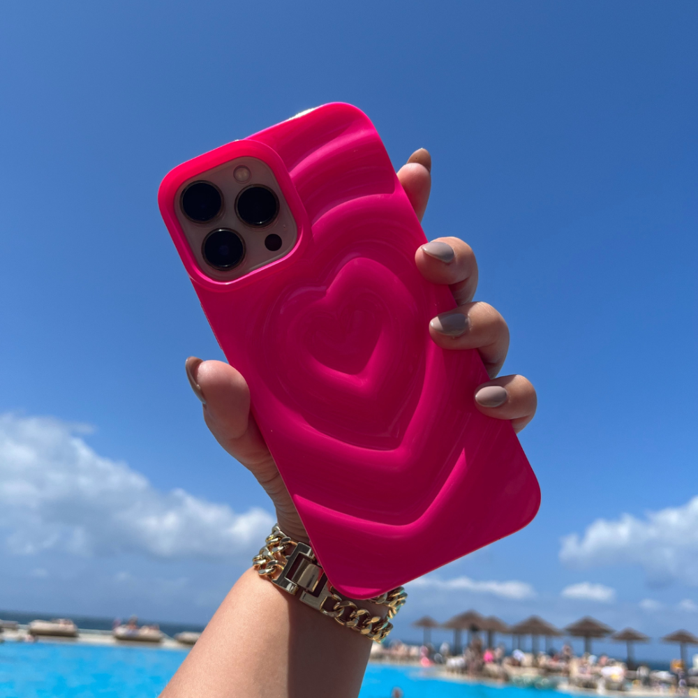 A hand adorned with a gold watch holds the Coconut Lane Melting Heart Phone Case in Bright Pink, showcasing its unique textured 3D melting heart design. The background reveals a clear blue sky dotted with scattered clouds, beach umbrellas, and a shimmering pool.