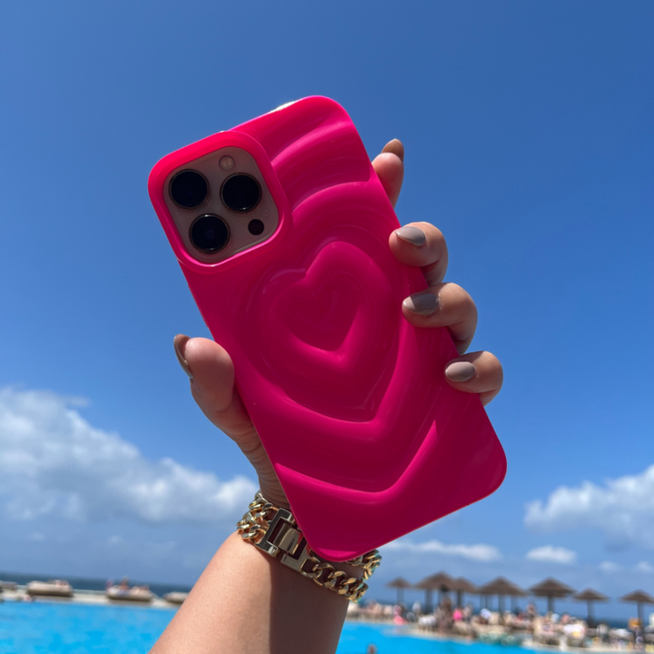 A hand adorned with a gold watch holds the Coconut Lane Melting Heart Phone Case in Bright Pink, showcasing its unique textured 3D melting heart design. The background reveals a clear blue sky dotted with scattered clouds, beach umbrellas, and a shimmering pool.