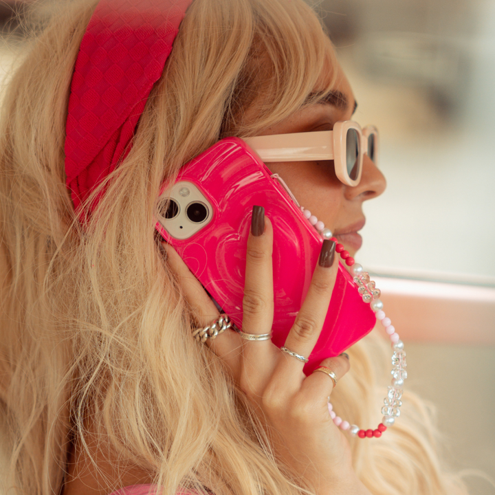 A person with long blonde hair is talking on a smartphone protected by Coconut Lane's Melting Heart Phone Case in bright pink. They wear white sunglasses, a pink headband, and multiple rings on their fingers. A beaded charm with pink and white beads hangs from the phone.