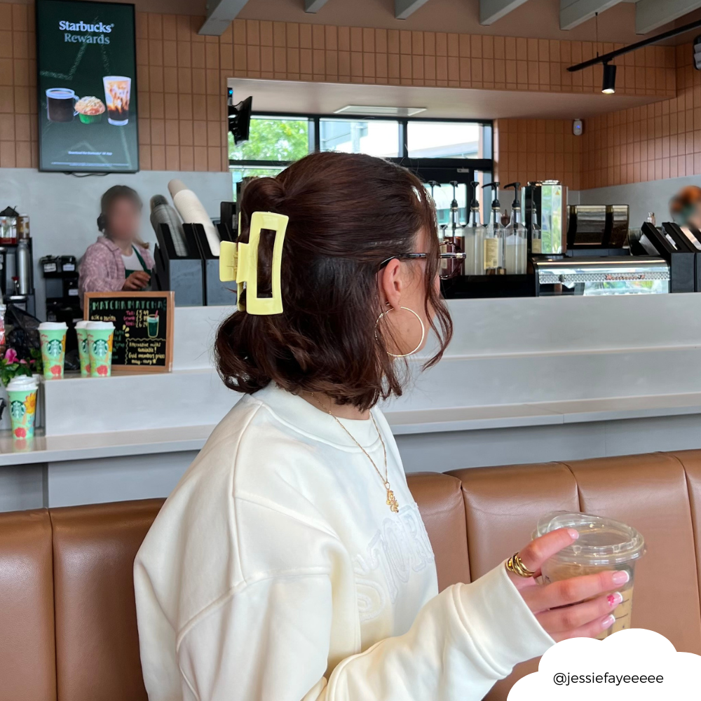 Woman with a yellow square Coconut Lane claw clip in her hair sits in Starbucks, holding a drink and facing away.