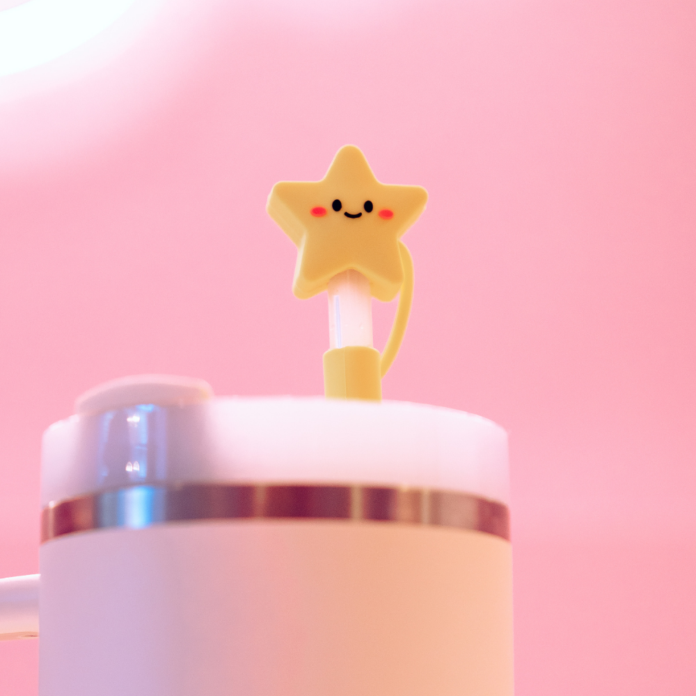 The Star Straw Cover by Coconut Lane is a delightful addition to a white tumbler with a pink lid. This adorable yellow silicone straw topper, shaped like a star, features a smiling face with rosy cheeks against a pink background, enhancing the cheerful and playful aesthetic of the set.