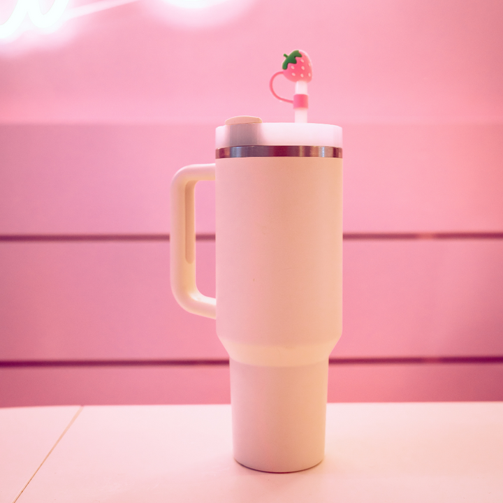 A tall, white travel mug with a handle sits on a pink surface. The mug features a lid adorned with Coconut Lane's Strawberry Straw Cover, and the background is pink, matching the overall aesthetic.