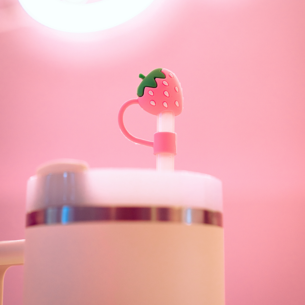 A white tumbler is adorned with a pink straw featuring Coconut Lane's Strawberry Straw Cover, a charming silicone topper shaped like a strawberry. The background is softly illuminated in pink hues by a circular light fixture, creating a cozy and playful atmosphere.
