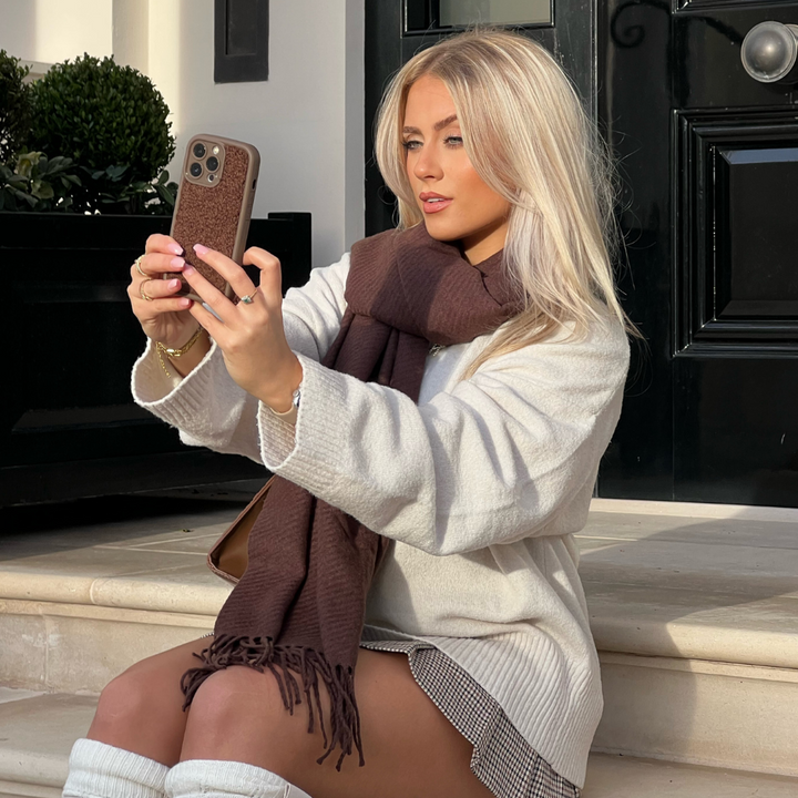 A blonde woman wearing a white sweater, brown scarf, and checkered skirt sits on steps taking a selfie with her smartphone encased in the Cosy Teddy Phone Case - Brown by Coconut Lane. She is outdoors in front of a black door and some potted plants, wearing knee-high white socks on a sunny day.