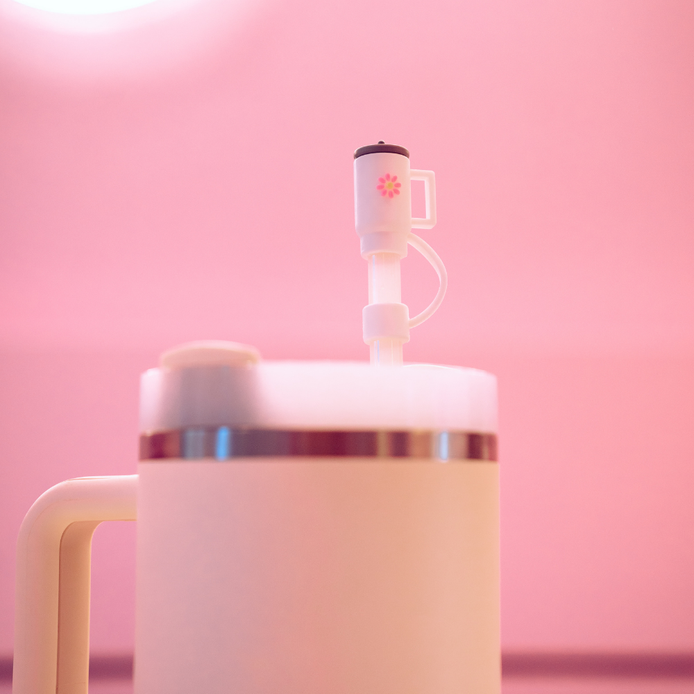 A close-up of a beige Coconut Lane Mini White Tumbler with a handle, containing a frothy beverage. A small device, possibly a portable milk frother or drink mixer, is inserted into the tumbler. The background is softly lit in a pink hue, and a Coconut Lane silicone Straw Cover lies nearby for added convenience and hygiene.