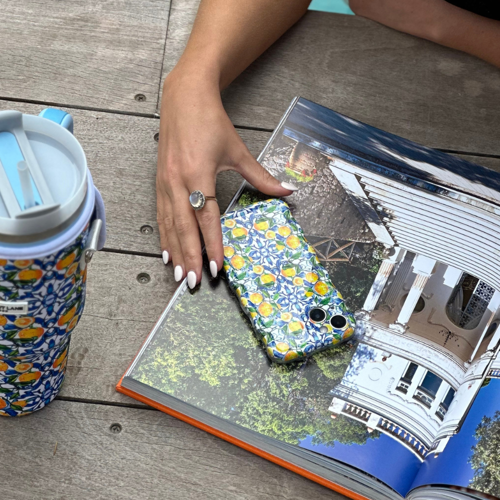 A person with white-painted nails and wearing a ring rests their hand on an open book featuring a coastal house. A phone in the Coconut Lane Wavy Phone Case - Amalfi Lemon lies on the book. Nearby, on the wooden surface, is a matching floral and Amalfi Lemon-patterned water bottle.