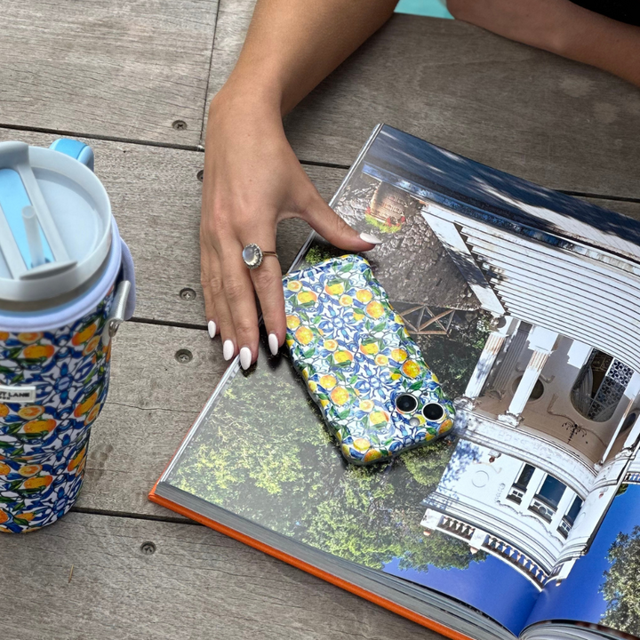 A person with white-painted nails and wearing a ring rests their hand on an open book featuring a coastal house. A phone in the Coconut Lane Wavy Phone Case - Amalfi Lemon lies on the book. Nearby, on the wooden surface, is a matching floral and Amalfi Lemon-patterned water bottle.
