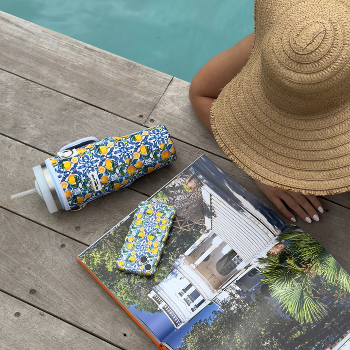 A person wearing a straw hat is lying beside a pool with an Amalfi Lemon-patterned tumbler, a Coconut Lane Wavy Phone Case - Amalfi Lemon, and a book featuring photographs of a house. The person has manicured white nails and is holding the book open to a page displaying an image of a white house.