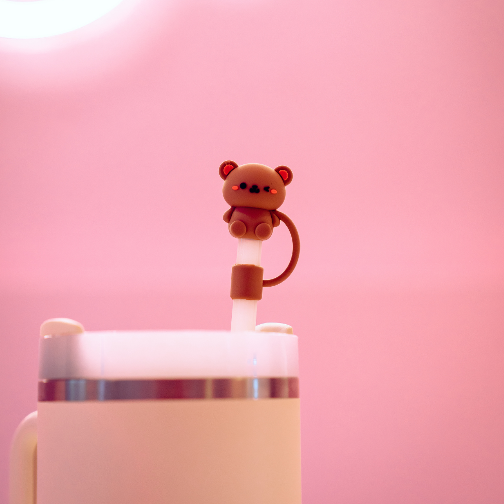 A Coconut Lane mug features a Teddy Bear Straw Cover, showcased against a soft pink background. The small, brown bear figure boasts charming red accents on its ears and cheeks. Complementing the design, the mug has a white body with a metallic band around the top.