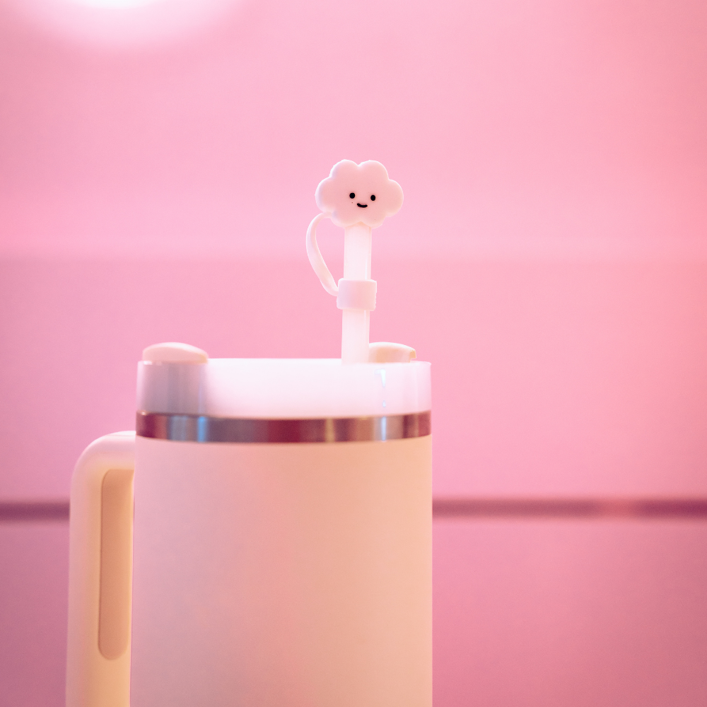 A white insulated travel mug with a handle on the left side is set against a pink background. The straw, fitted with Coconut Lane's White Cloud Straw Cover featuring a cute cloud-shaped silicone cover adorned with a smiling face, complements the design. The lid of the mug is secured with a stainless steel band.