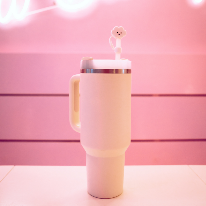A pink travel mug with a handle stands on a matching pink surface. The mug has a lid equipped with a metal band and a stirring rod topped with a small, smiling flower-shaped decoration. Adding to its charm, it's designed to complement accessories like Coconut Lane's White Cloud Straw Cover for convenience. The background features a gradient of pink shades and a neon light.