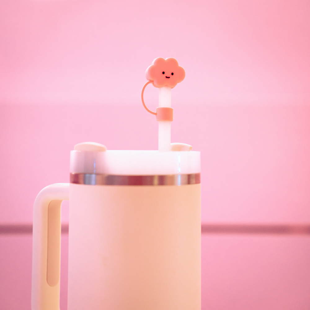 A close-up of a white travel mug with a handle and a stainless steel rim against a pink background. The straw showcases Coconut Lane's Pink Cloud Straw Cover, featuring an adorable smiling pink cloud-shaped silicone cover, adding a playful touch to the design that's both functional and charming.