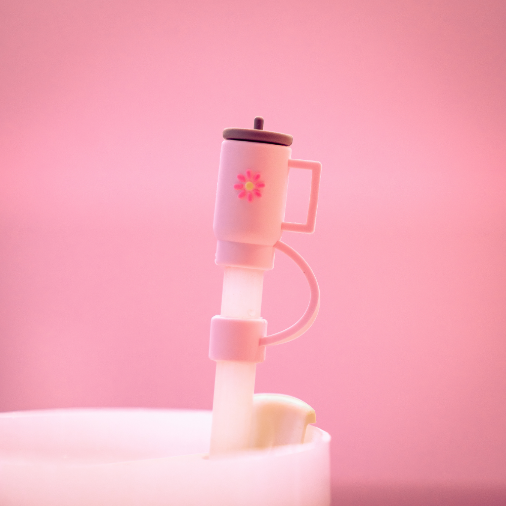 The Mini Pink Tumbler Straw Cover by Coconut Lane features a small, pink plastic coffee cup charm adorned with a tiny flower design, attached to a translucent silicone straw topper. The cohesive pastel aesthetic is complemented by a soft pink background.